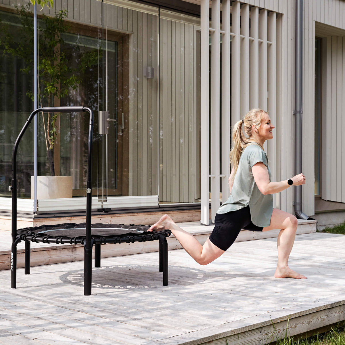 A woman is doing bending exercises on an Acon FIT Balance rebounder on a house terrace.