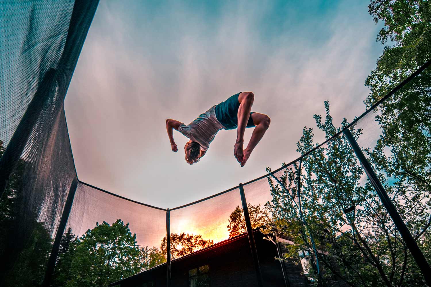 A boy in blue shorts doing a backflip on a trampoline in a sunset.