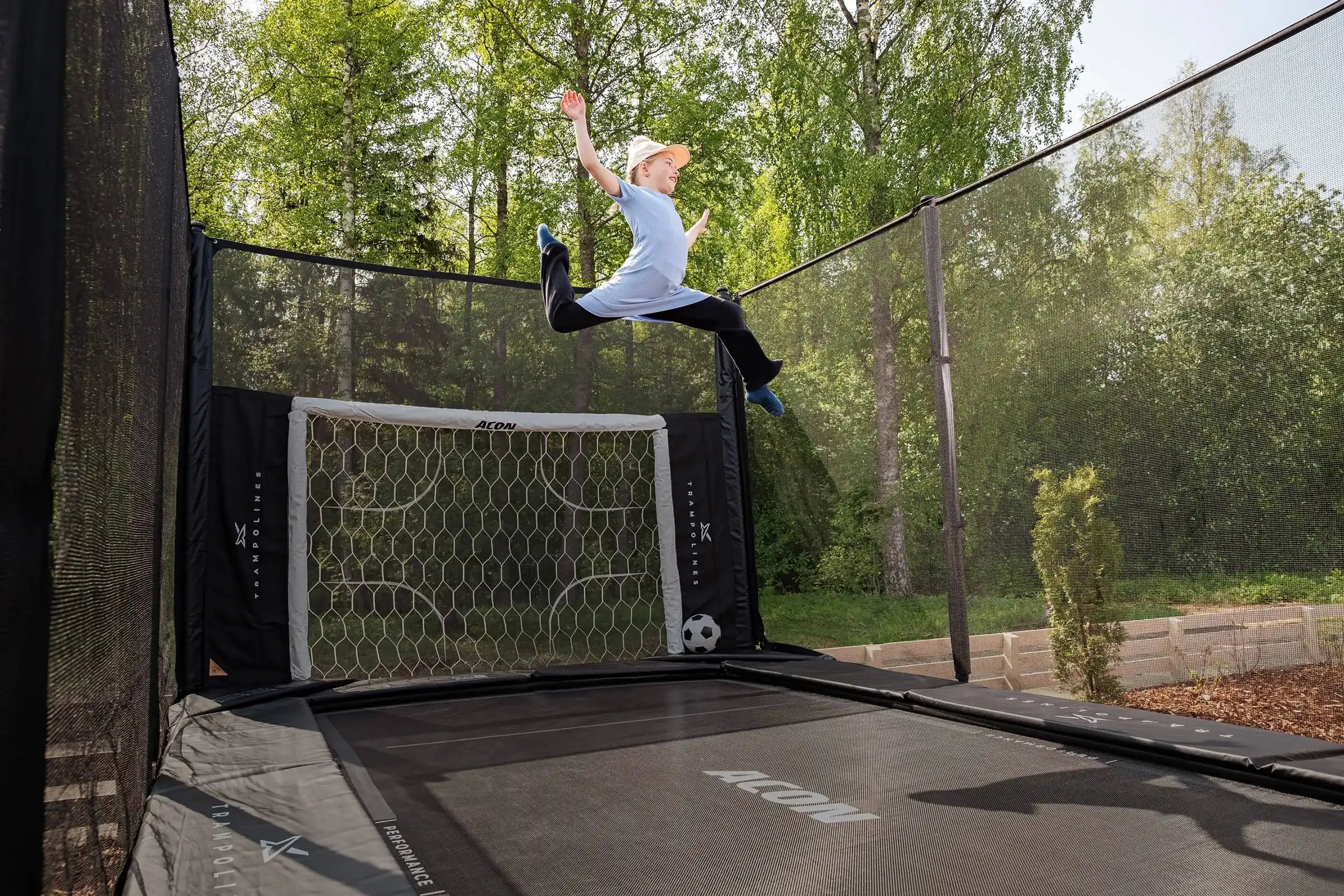 Girl jumping on an Acon X trampoline in a sunny backyard, equipped with an Acon soccer goal.