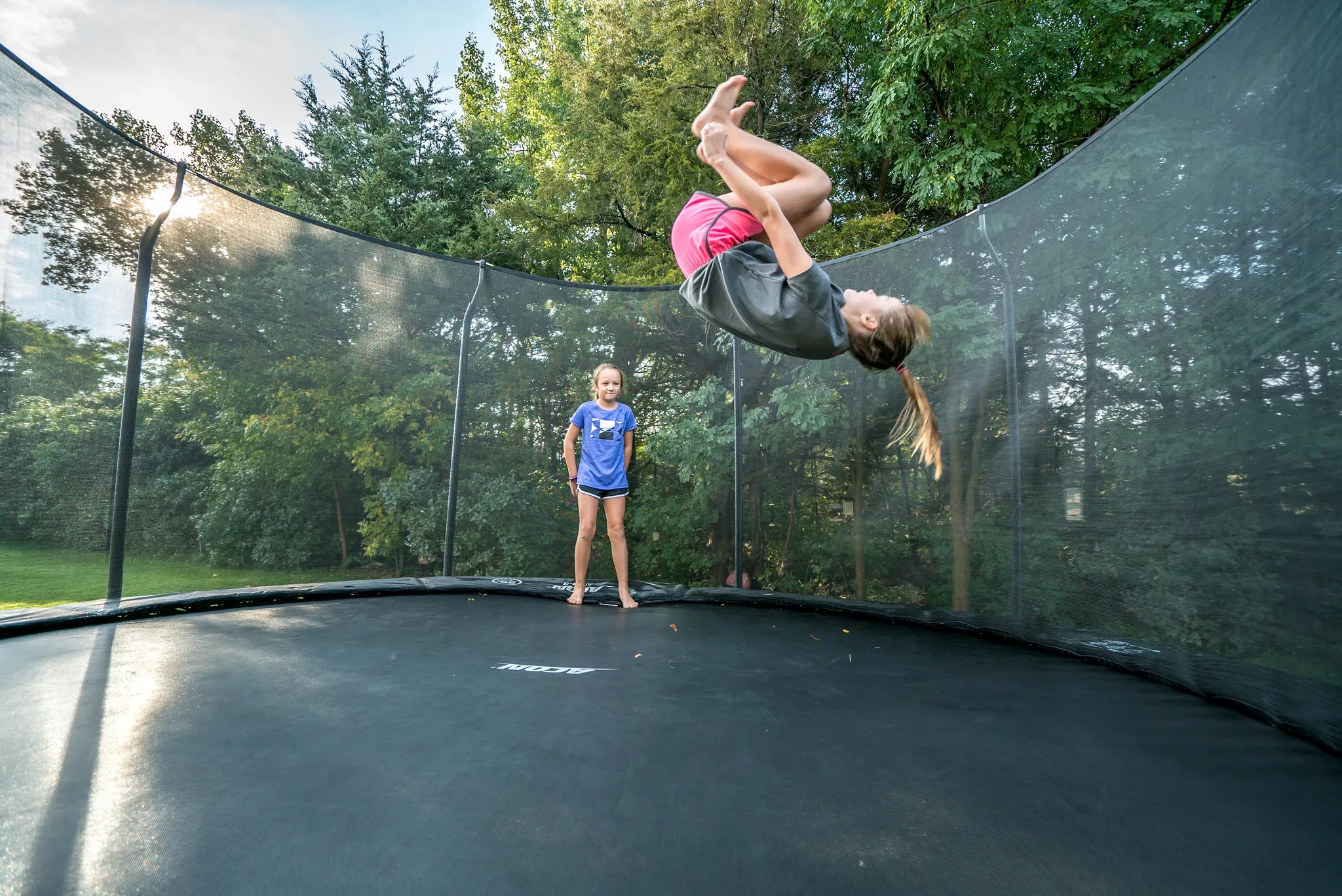 Two girls inside of round Acon trampoline with safety net, other is performing a backflip while other is watching 