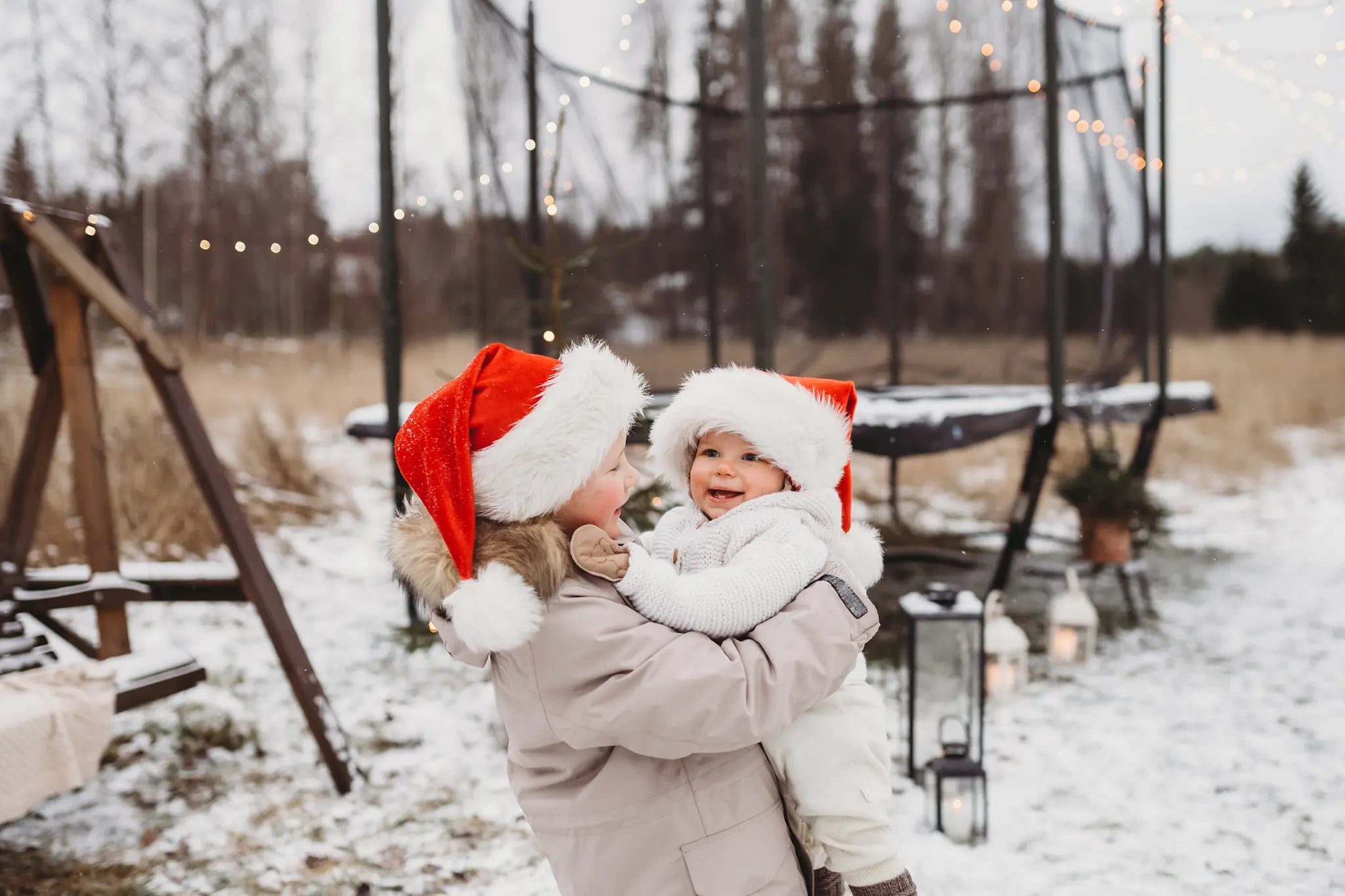 Two children wearing Santa hats smile and hug outdoors in a snowy yard, with a decorated trampoline and string lights in the background.