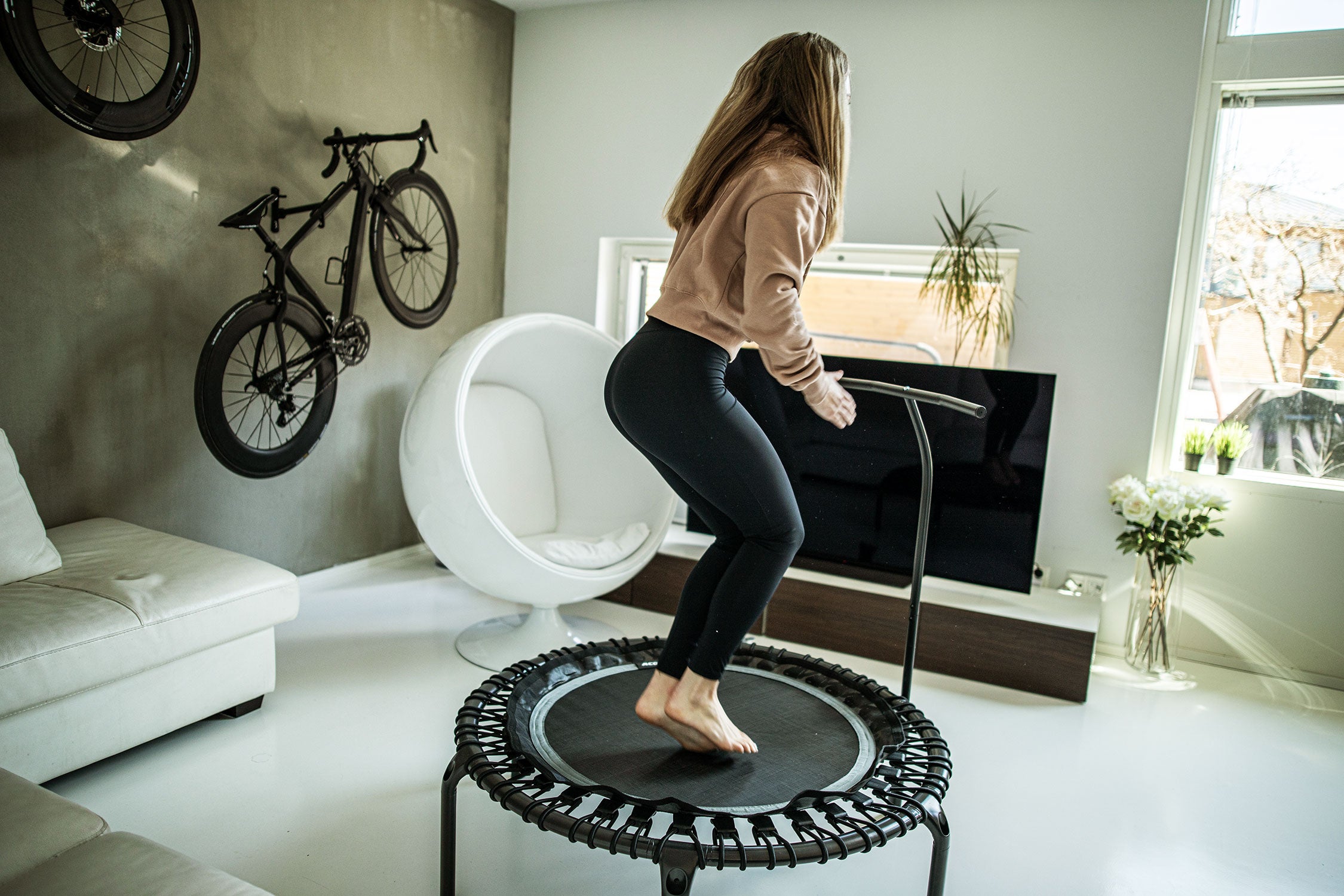 A woman doing ski jumps on a black and round rebounder trampoline at her home in her living room