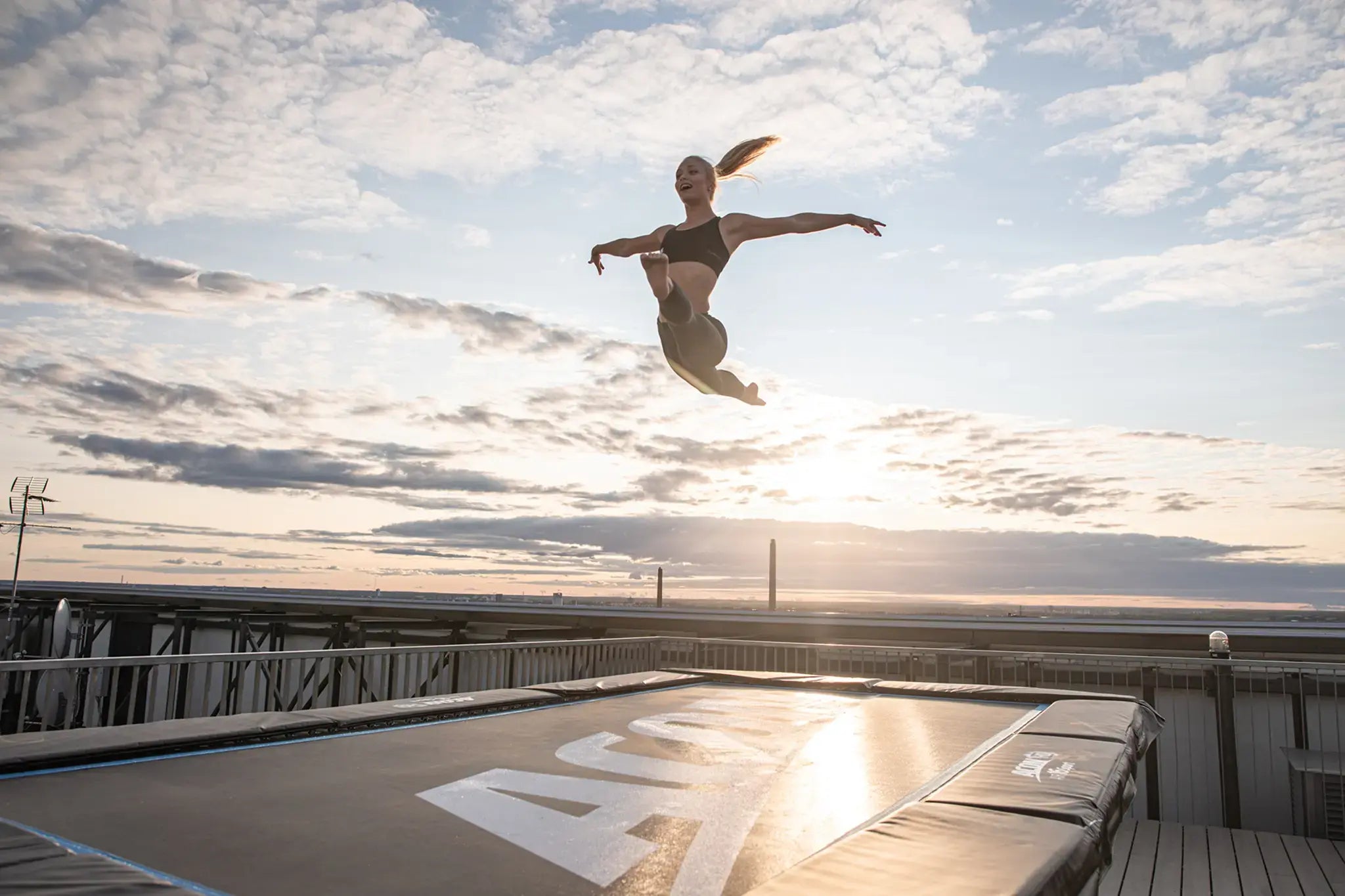 Person performing a high jump on an Acon trampoline at sunset, with a cityscape and clouds in the background.