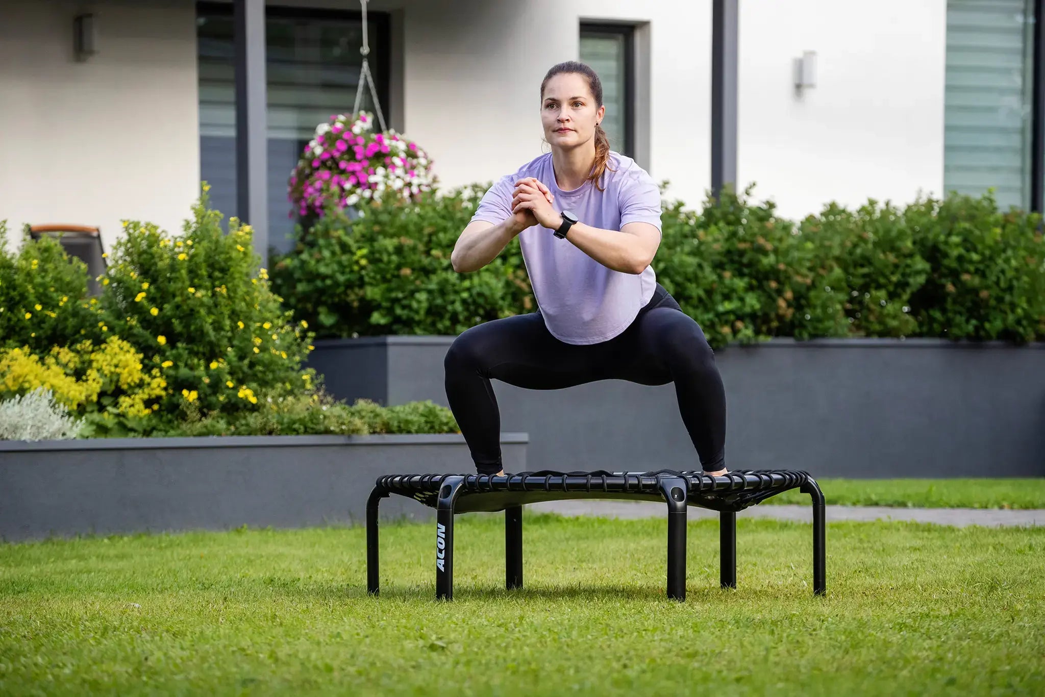 Woman performs a squat on an ACON rebounder trampoline in a backyard, wearing sports clothing and a smartwatch, with greenery and flowers in the background.