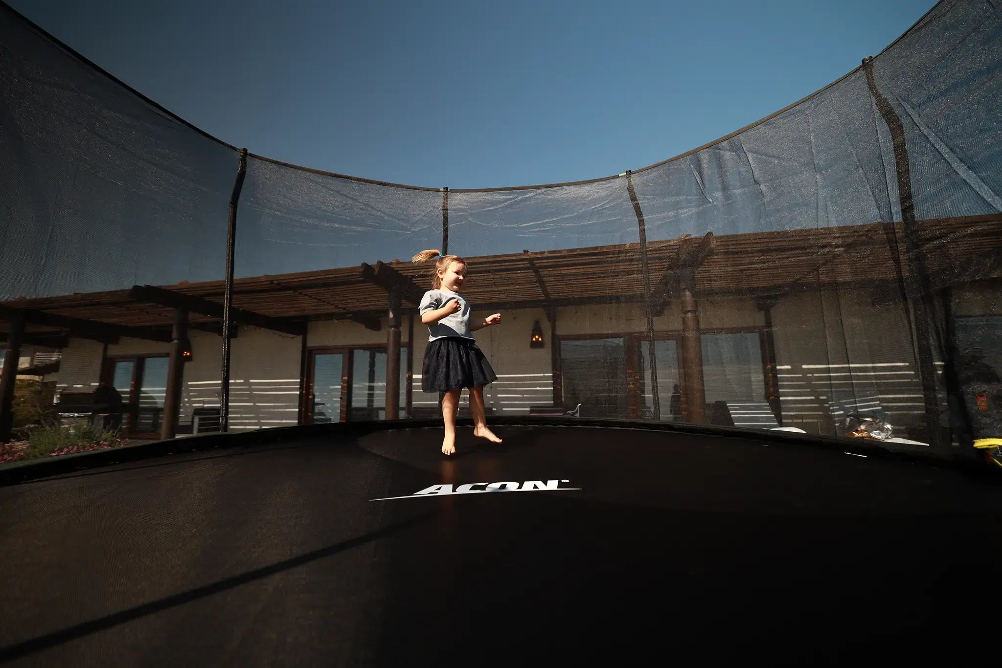 A young girl jumping on a ACON trampoline in a backyard with a house in the background.