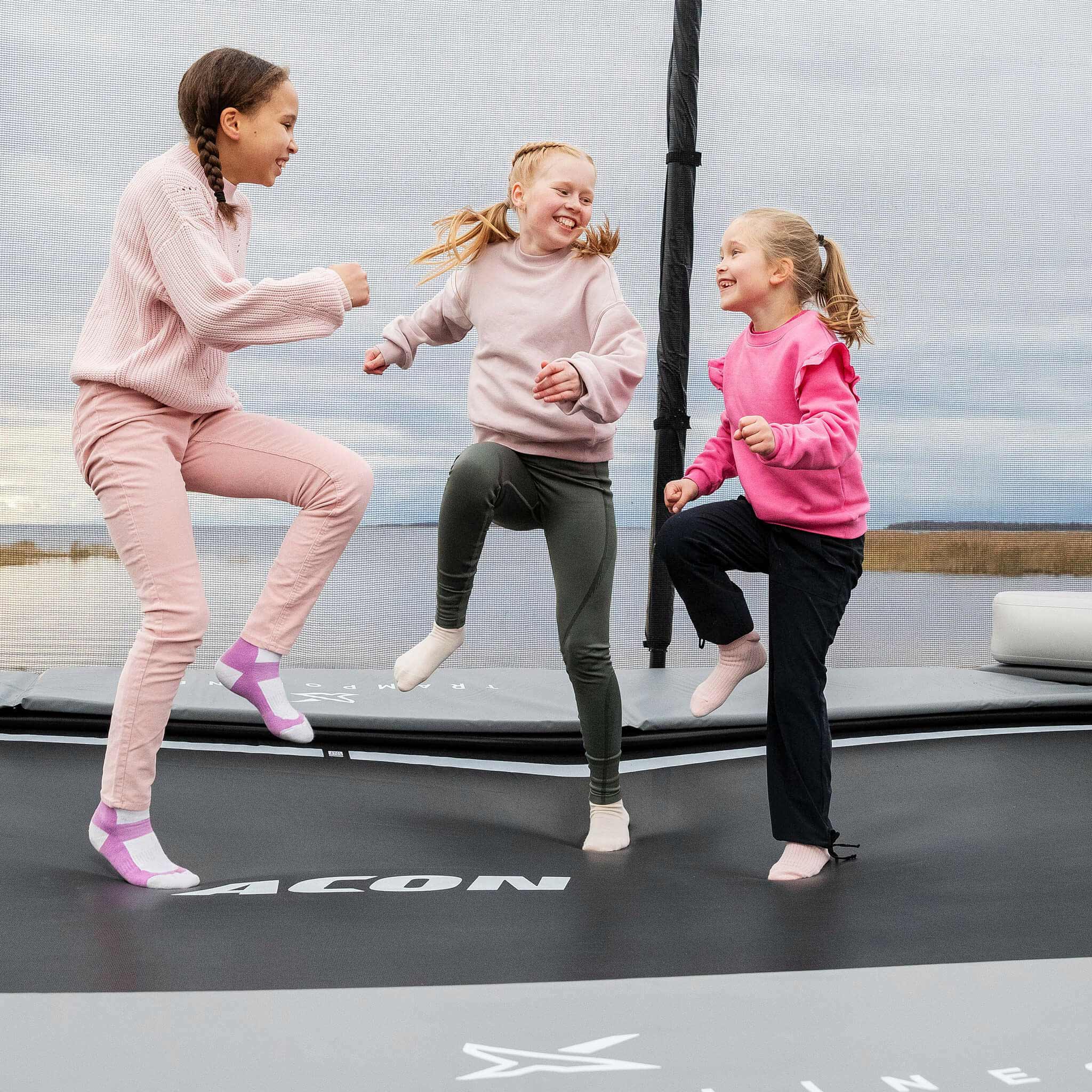 Three happy girls in pink shirts step on the Acon X Trampoline.